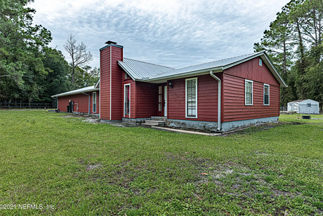 Red-sided house with a metal roof and chimney on a grassy lot, surrounded by trees under a cloudy sky.