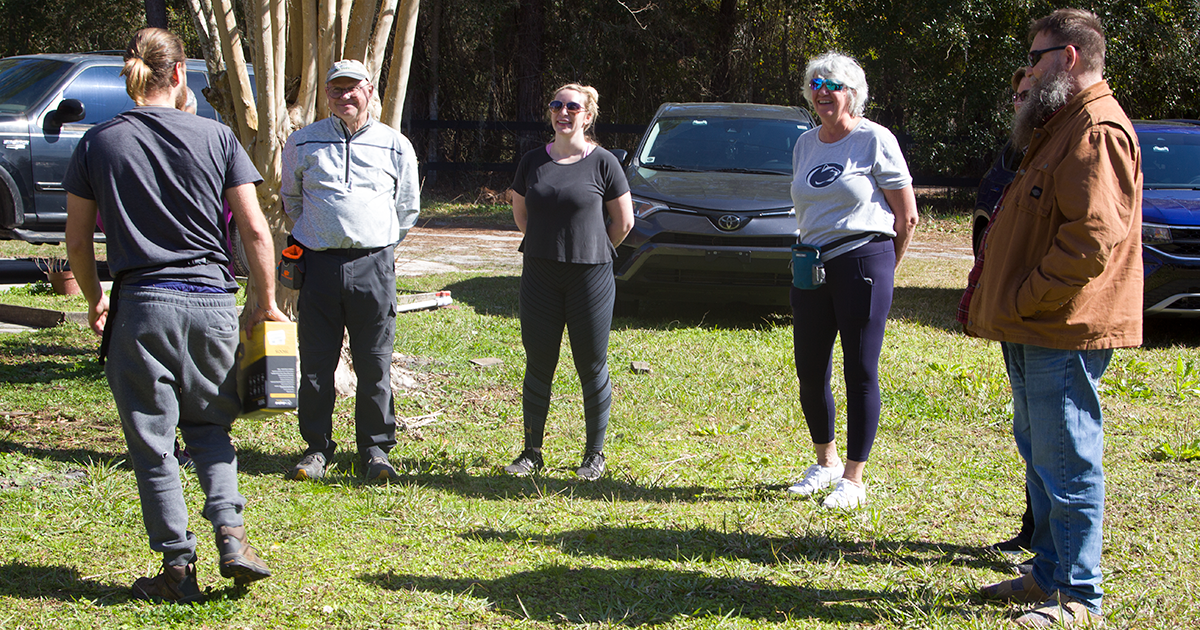 A group of six people standing in a circle outdoors. Some are looking at each other, others at the ground.