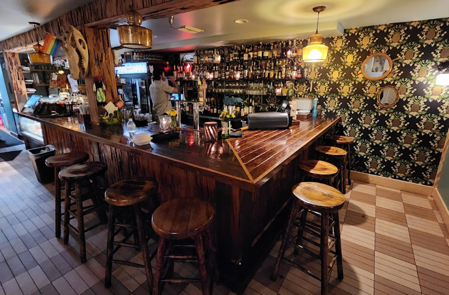 Bar interior with wooden counter and stools, bartender, and patterned wallpaper.