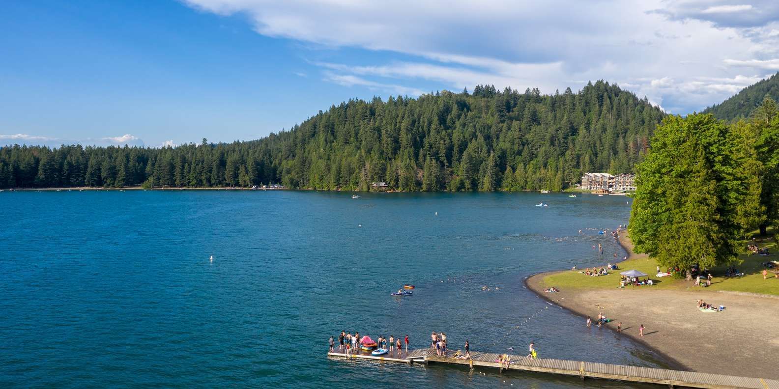Cultus lake surrounded by trees with a beach, and people. Blue water under a partly cloudy sky.
