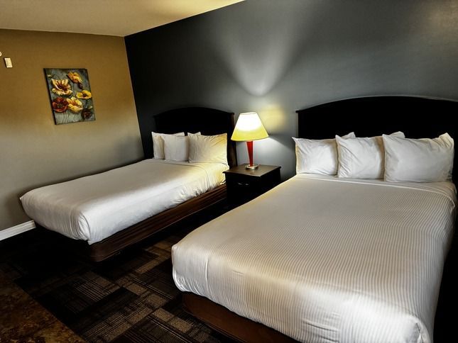 Two beds in a hotel room with white linens, lamp on nightstand, art on the wall.