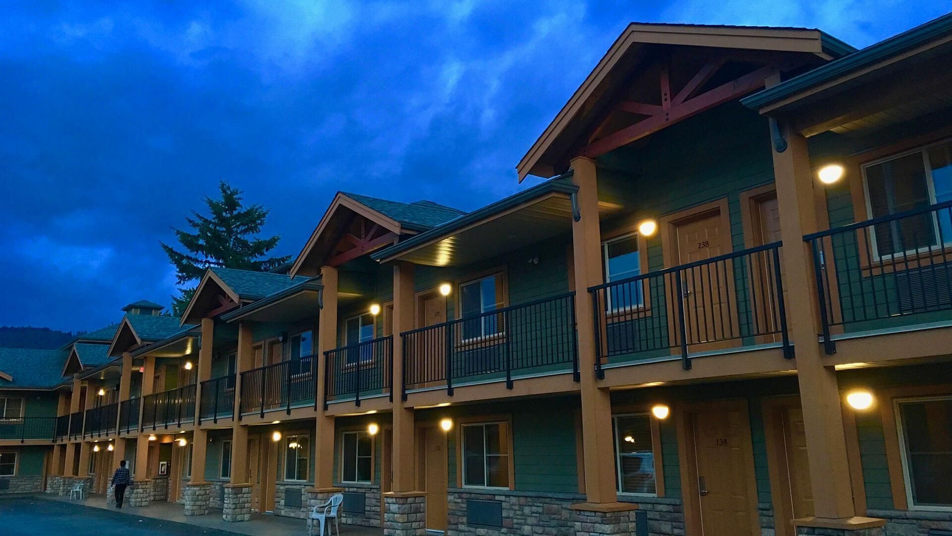 Motel exterior at dusk with green siding, wooden accents, and lit doorways.