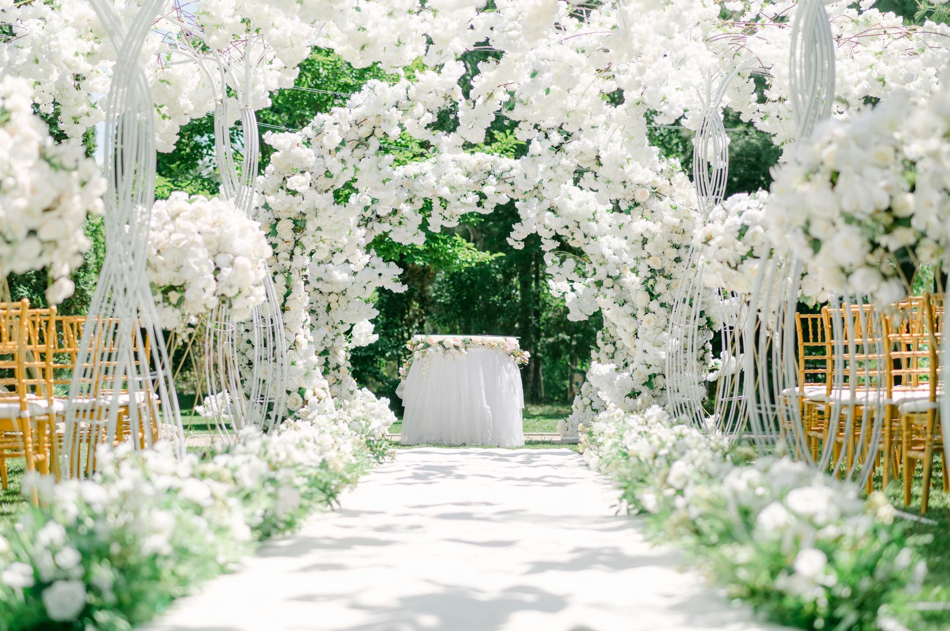 Wedding ceremony setup with white floral arch, aisle, and gold chairs in a garden.