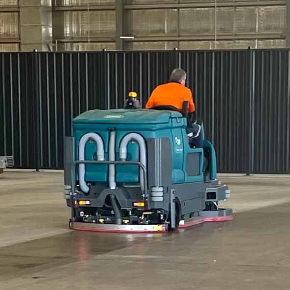 Man in orange shirt operating a teal floor scrubber in a large warehouse — Industrial Power Sweeping Services Pty Ltd In Knuckey Lagoon, NT