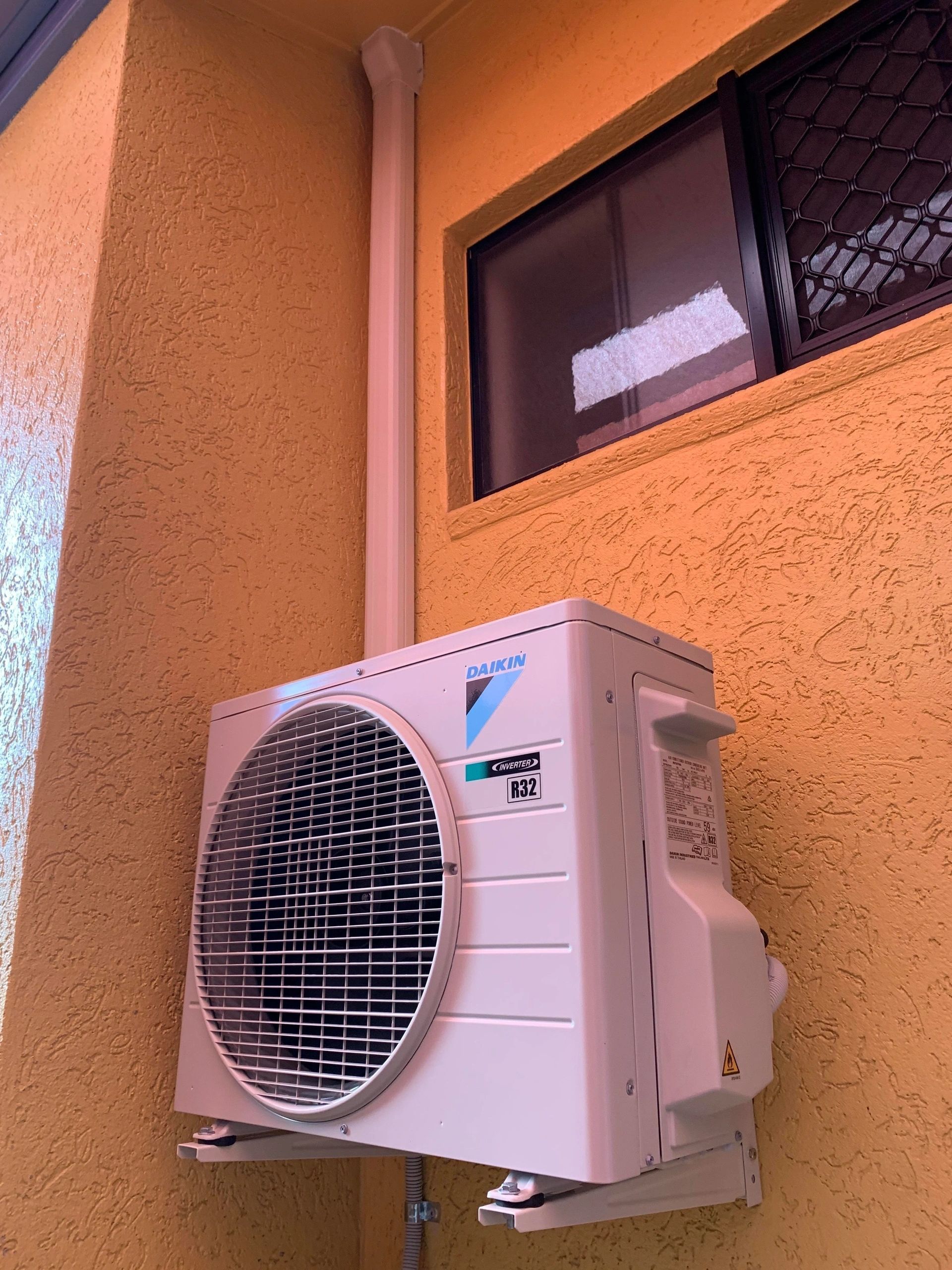 A White Air Conditioner Is Mounted On A Yellow Wall Next To A Window  — Law Air Conditioning In Ingham, QLD