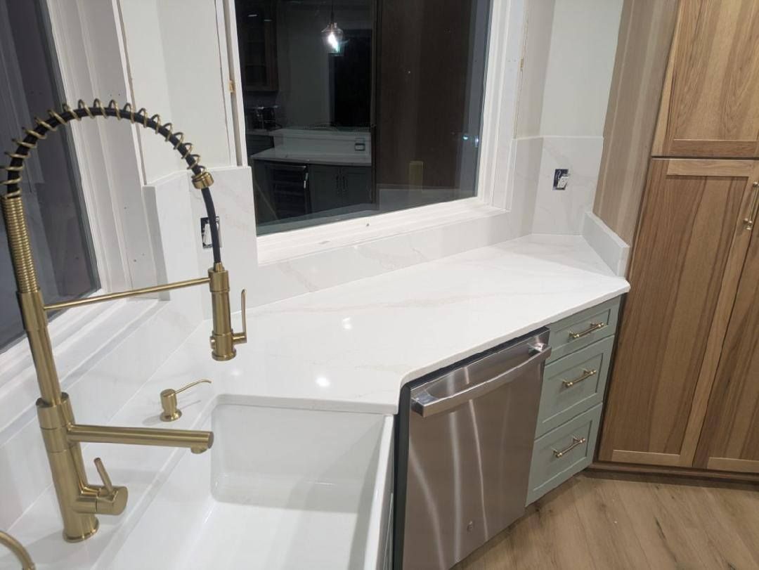 Modern kitchen sink with gold faucet, white countertop, window, and stainless dishwasher.
