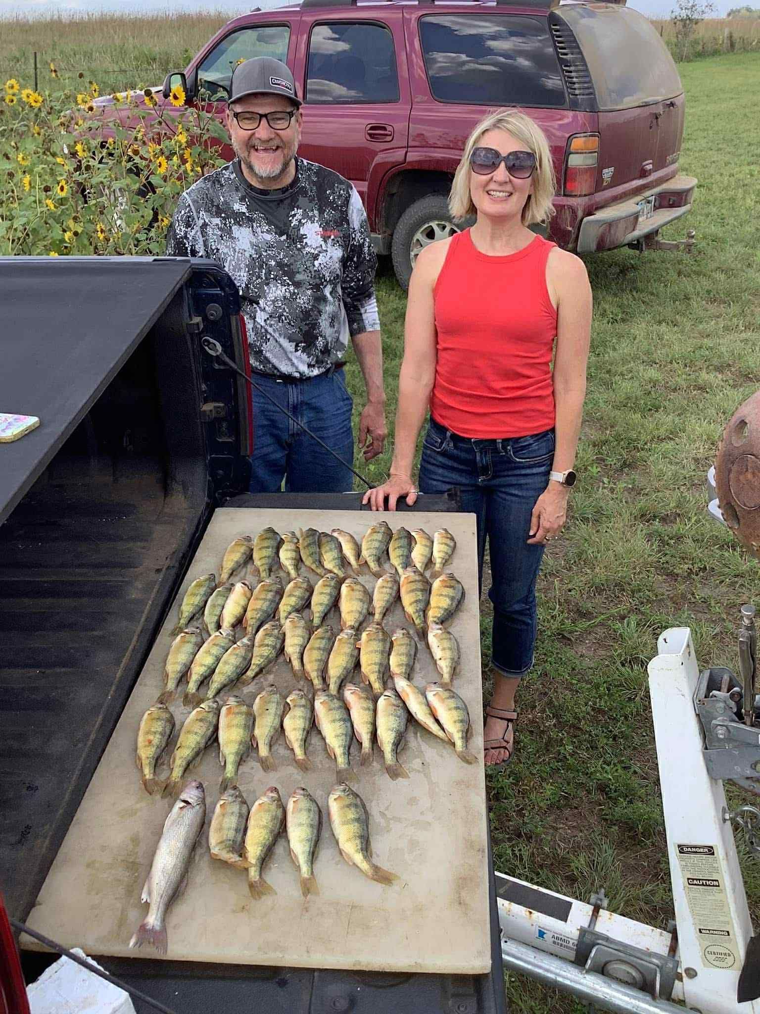 Man and woman beside a table displaying many fish; truck in background; outdoors.