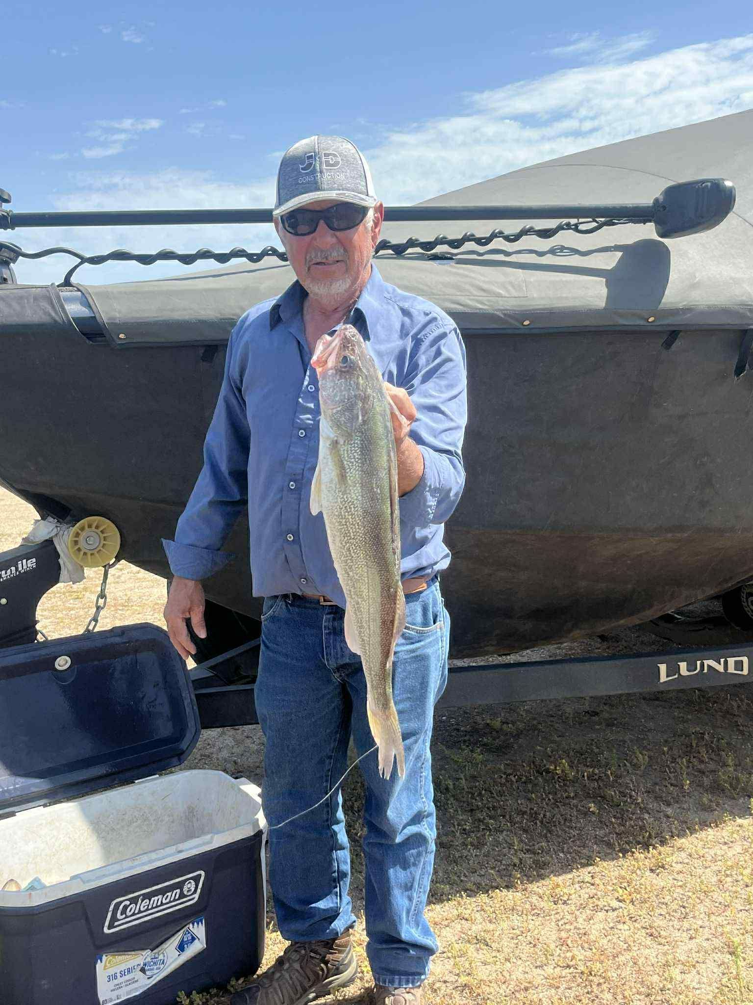 Man holds a fish, standing near a boat. He is wearing a blue shirt, jeans, and a cap.