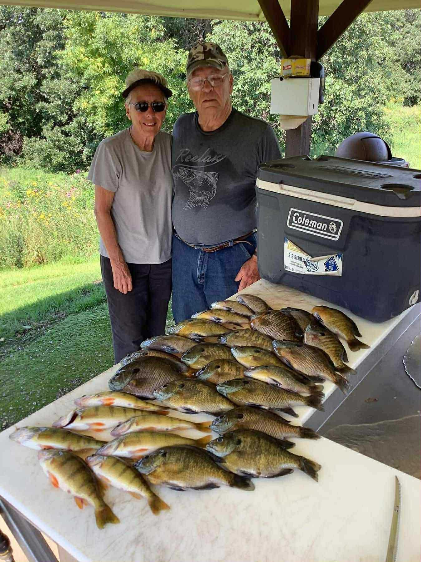 Two people pose with many fish on a table; outdoors, sunny. Coleman cooler on table.