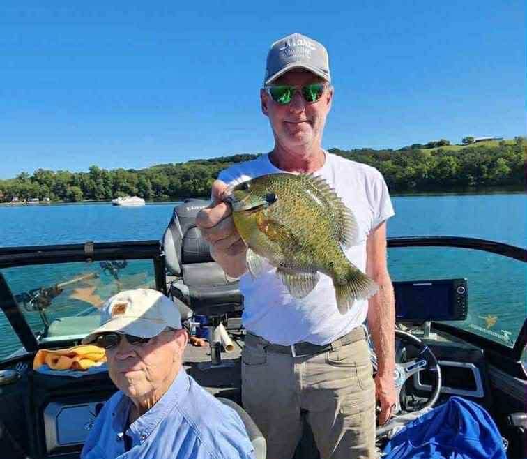 Man on a boat holding a large sunfish; another man seated nearby. Outdoors, sunny.