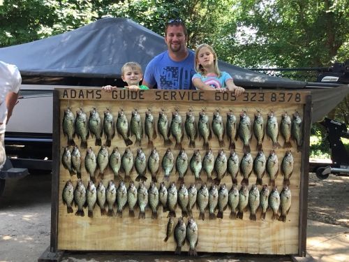A man and two children pose with a large display of caught fish in front of a boat.