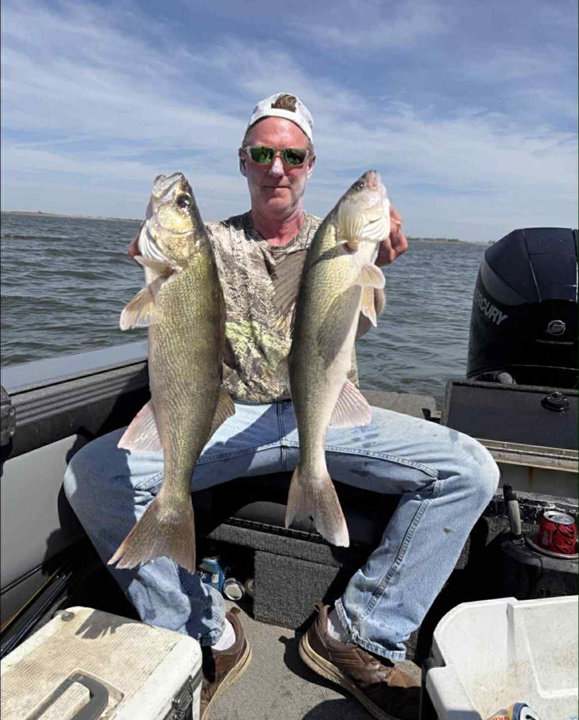 Man on a boat holds up two walleye fish. Gray and tan fish. Water and sky in background.