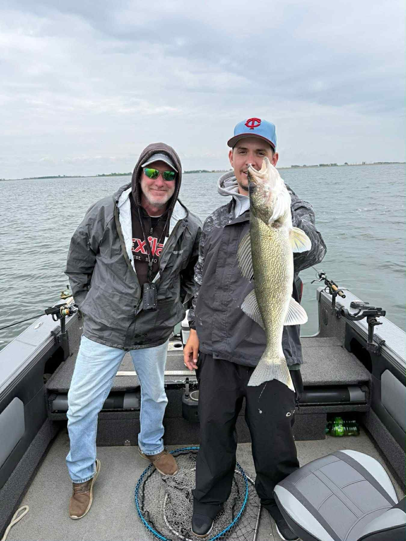 Two men on a boat, one holding a large fish. Overcast sky, water visible.