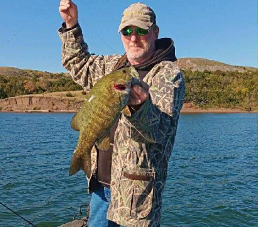 Man in camo holding a large fish, on a boat near a lake, under a blue sky.