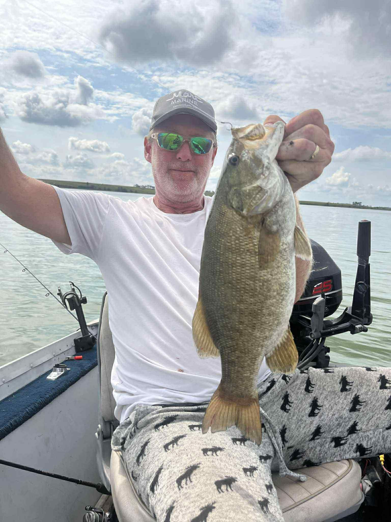 Man on boat holding up a fish, cloudy day, water in background.