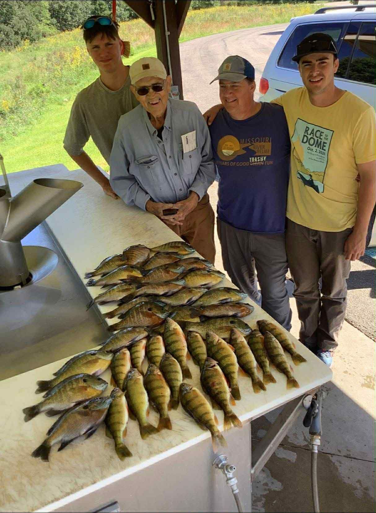 Four people pose with a large catch of yellow perch and bluegill on a white table under a shelter.