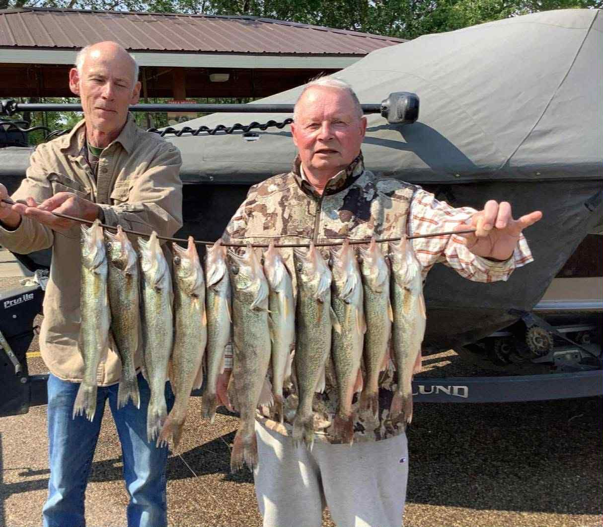 Two men holding a string of caught fish near a boat. One wears camouflage, the other a light brown shirt.
