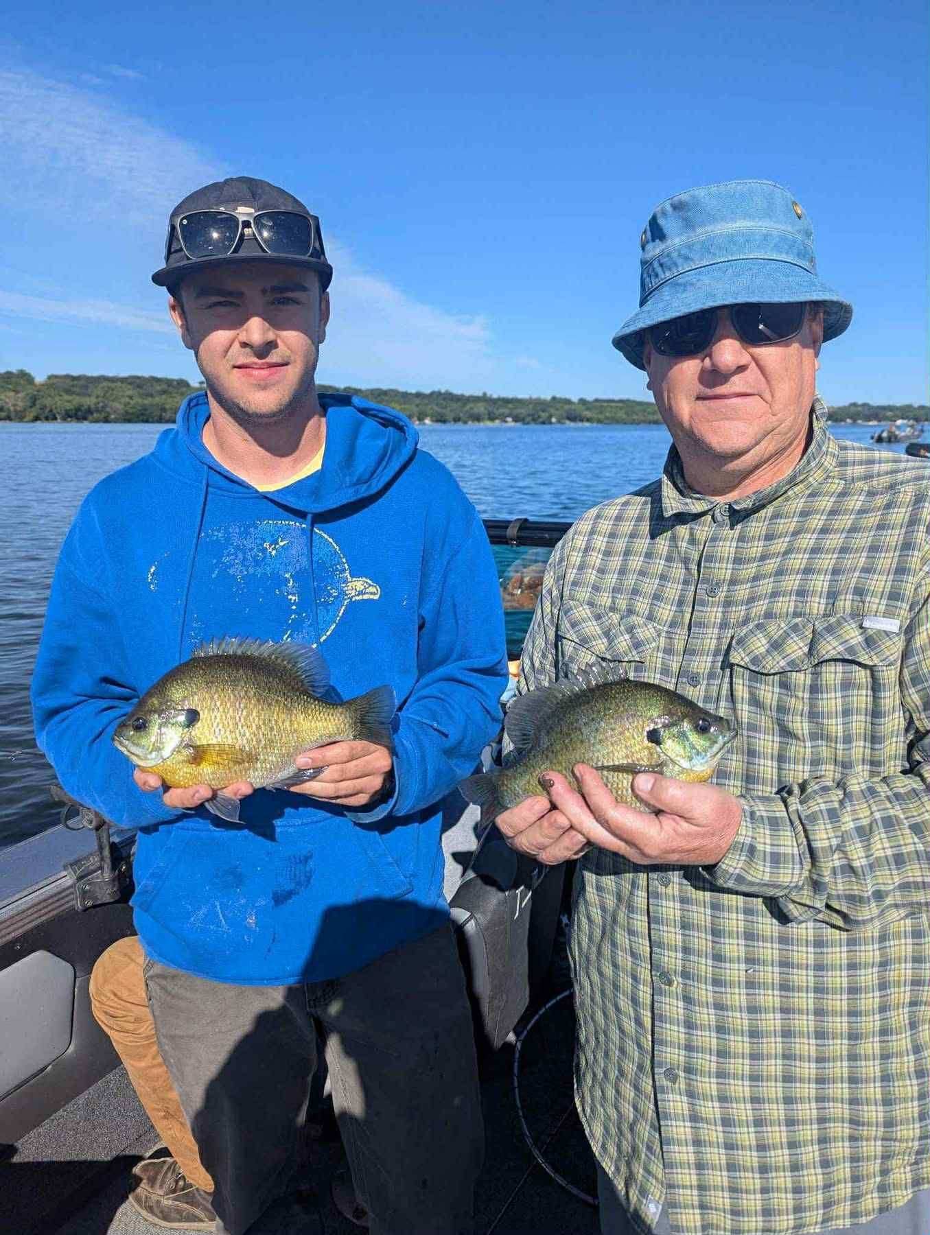 Two people holding up fish on a boat. One wears a blue hoodie, the other a plaid shirt. Clear sky, lake.