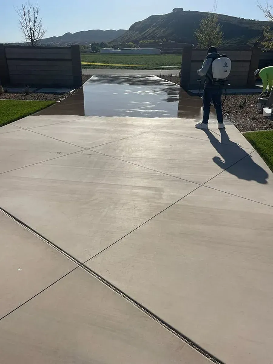 Man spraying a driveway with a backpack sprayer, concrete, with a mountain backdrop.