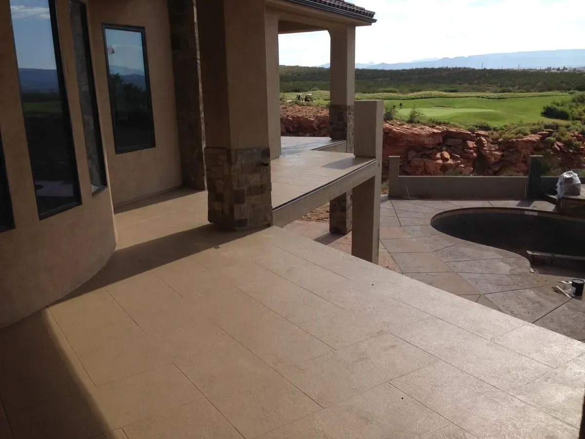 Beige patio overlooking a green landscape with a pool and windows.