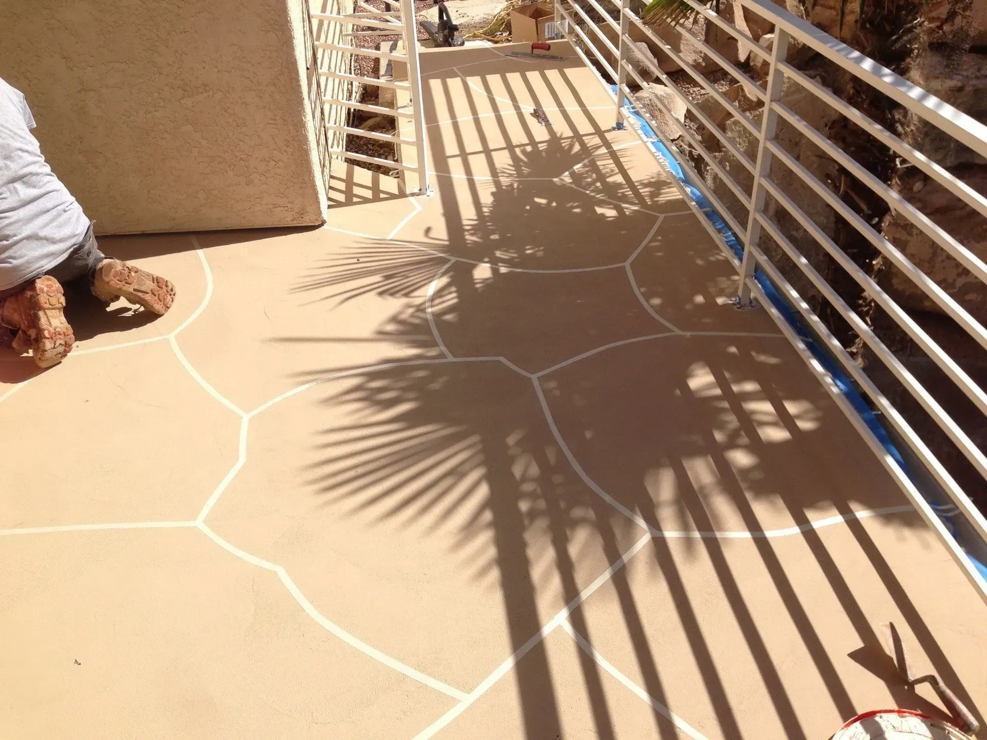A person applying white lines to a tan, decorative concrete surface. Shadows of a palm tree and railing.