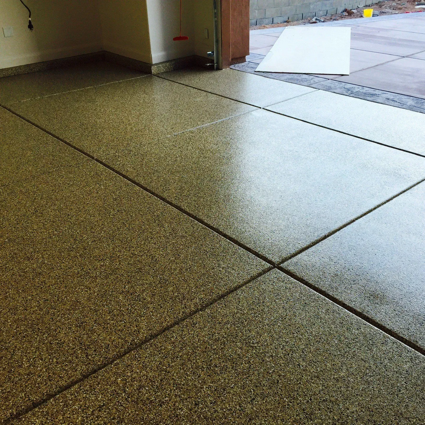 Epoxy-coated garage floor with tan and brown flecks, showing control joints and a partially open garage door.