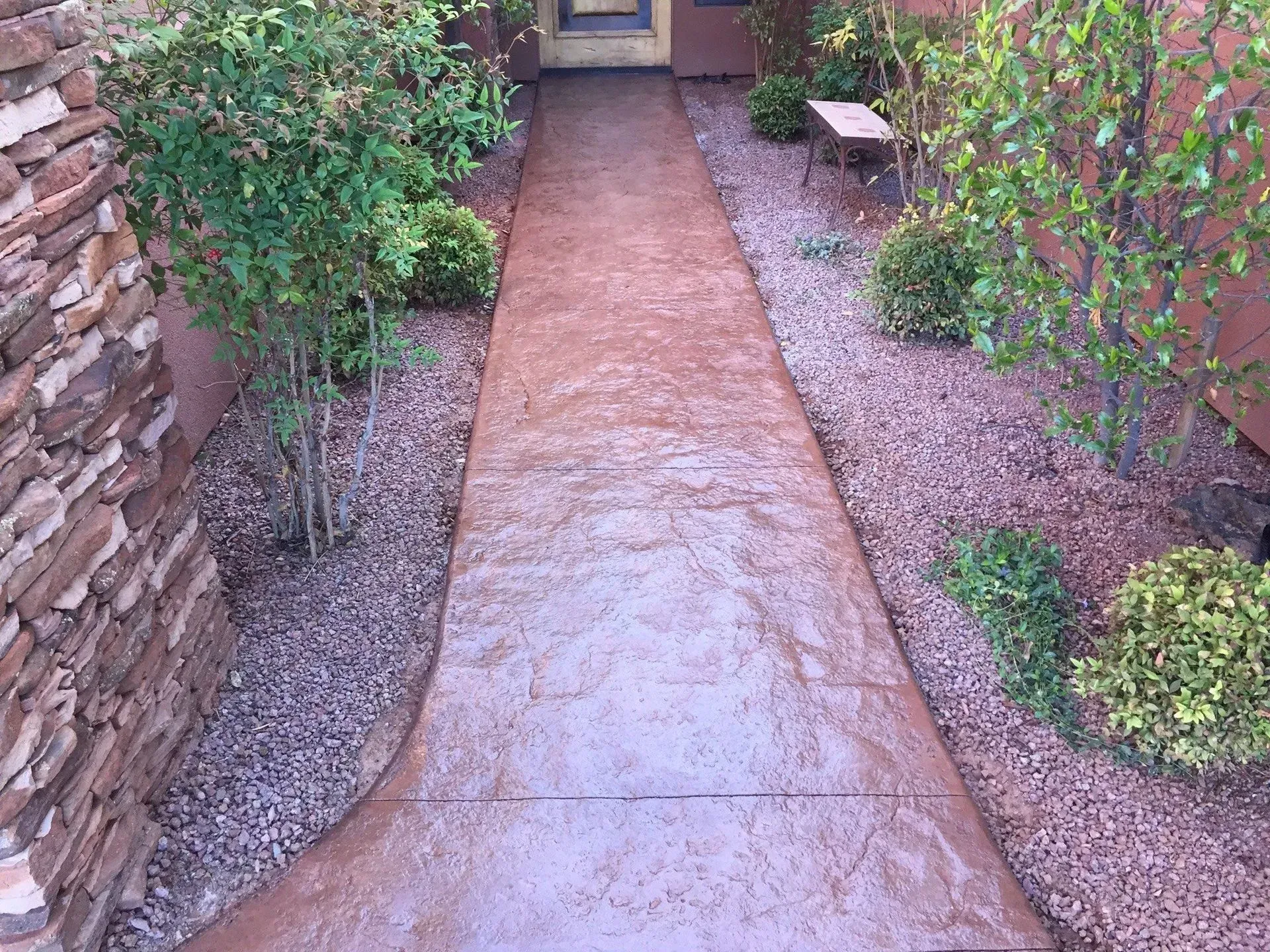 A brown stamped concrete walkway flanked by bushes, gravel, and a stone wall.