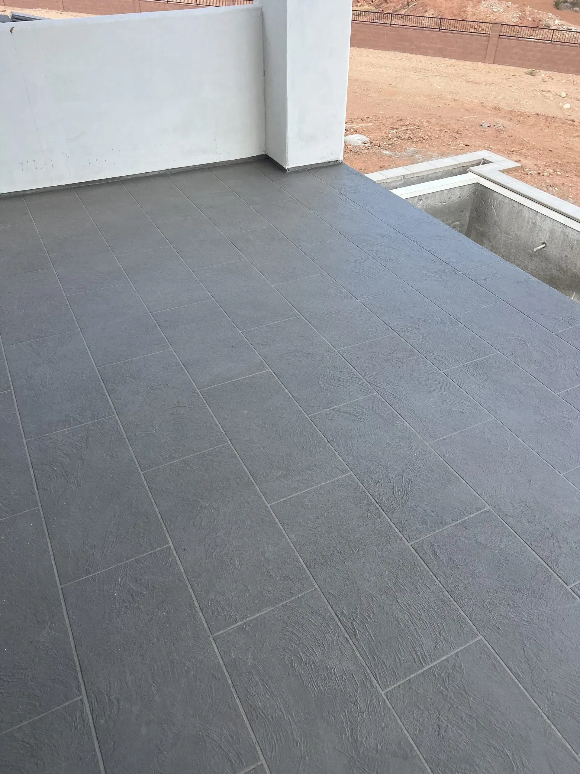 Gray tiled outdoor floor, next to a concrete wall and a drainage area, viewed from above.