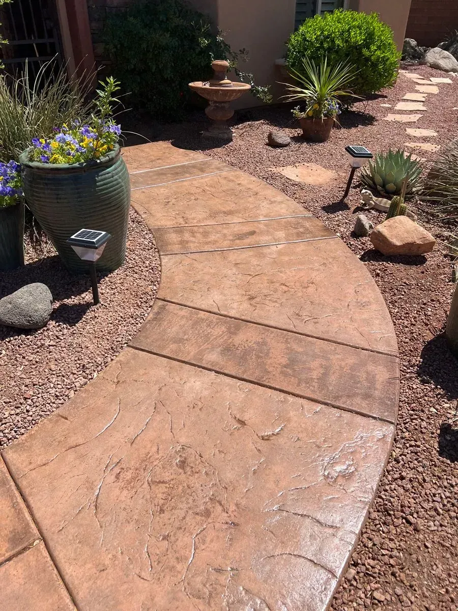 Curved walkway with textured concrete, surrounded by red gravel, leading to a garden with plants and a fountain.