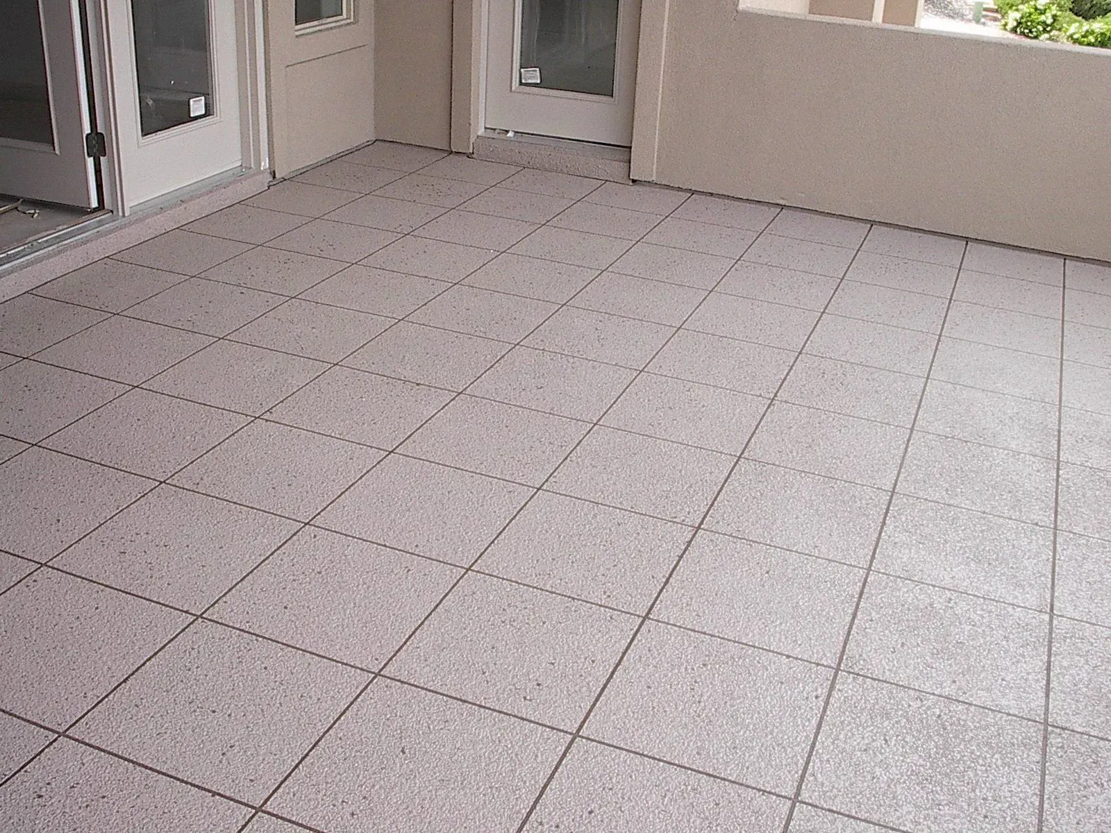 Tiled patio with light gray tiles and gray grout. Two sets of glass doors and light-colored walls are present.