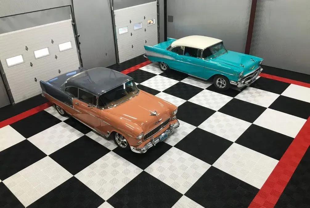 Two old cars are parked on a checkered floor in a garage.