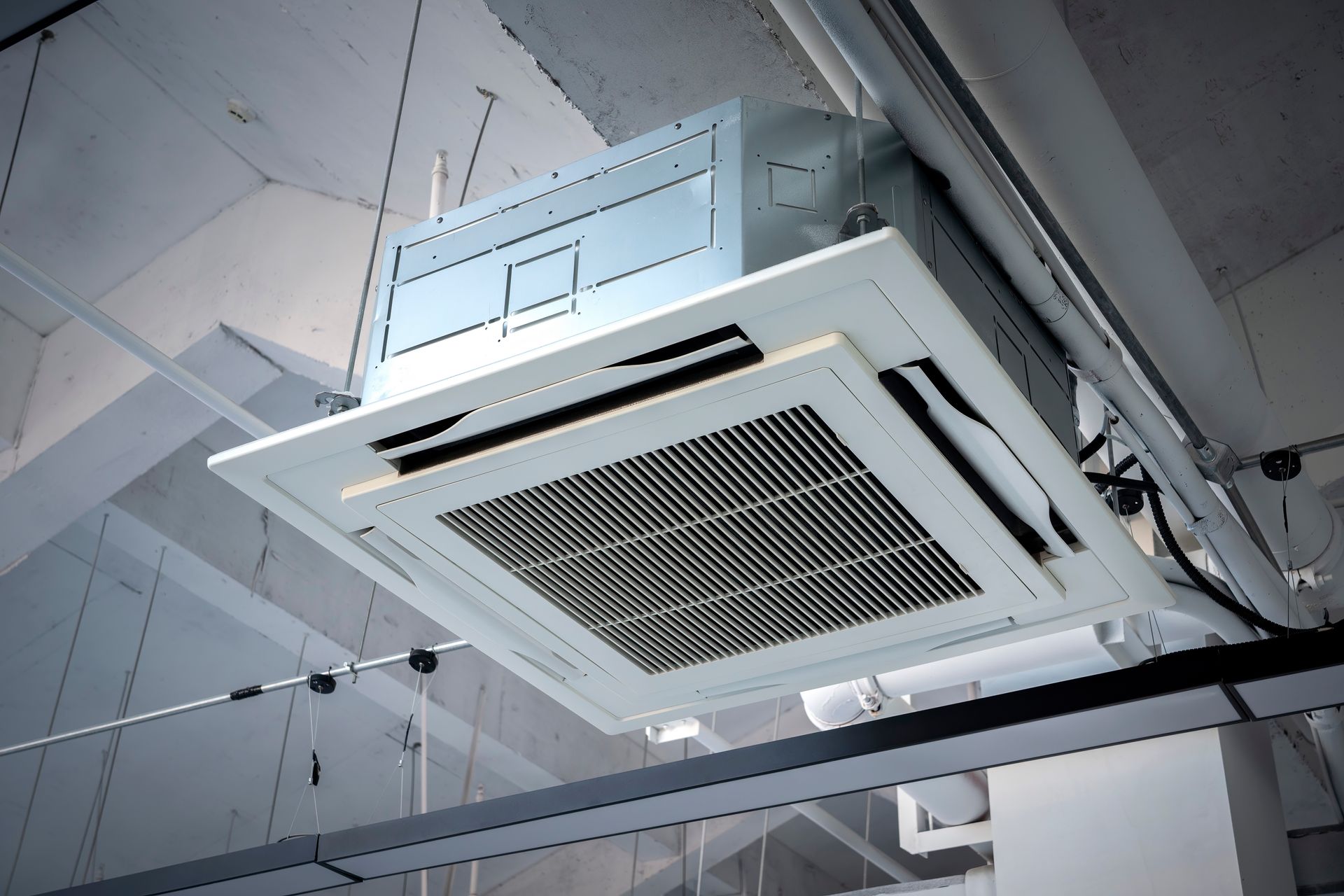 Ceiling-mounted air conditioning unit with square vent, white frame, suspended in an indoor setting.