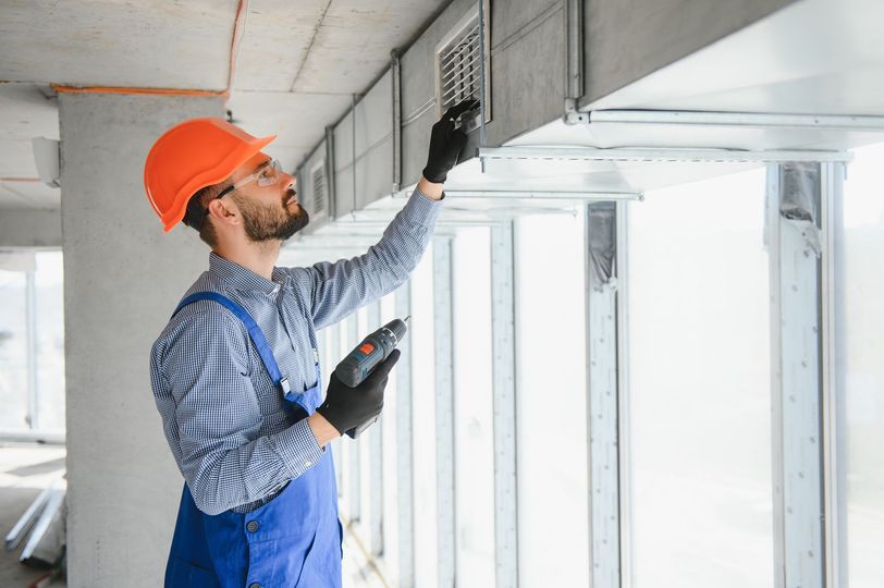 HVAC worker in orange hard hat and blue overalls inspecting a vent. Unfinished building interior.
