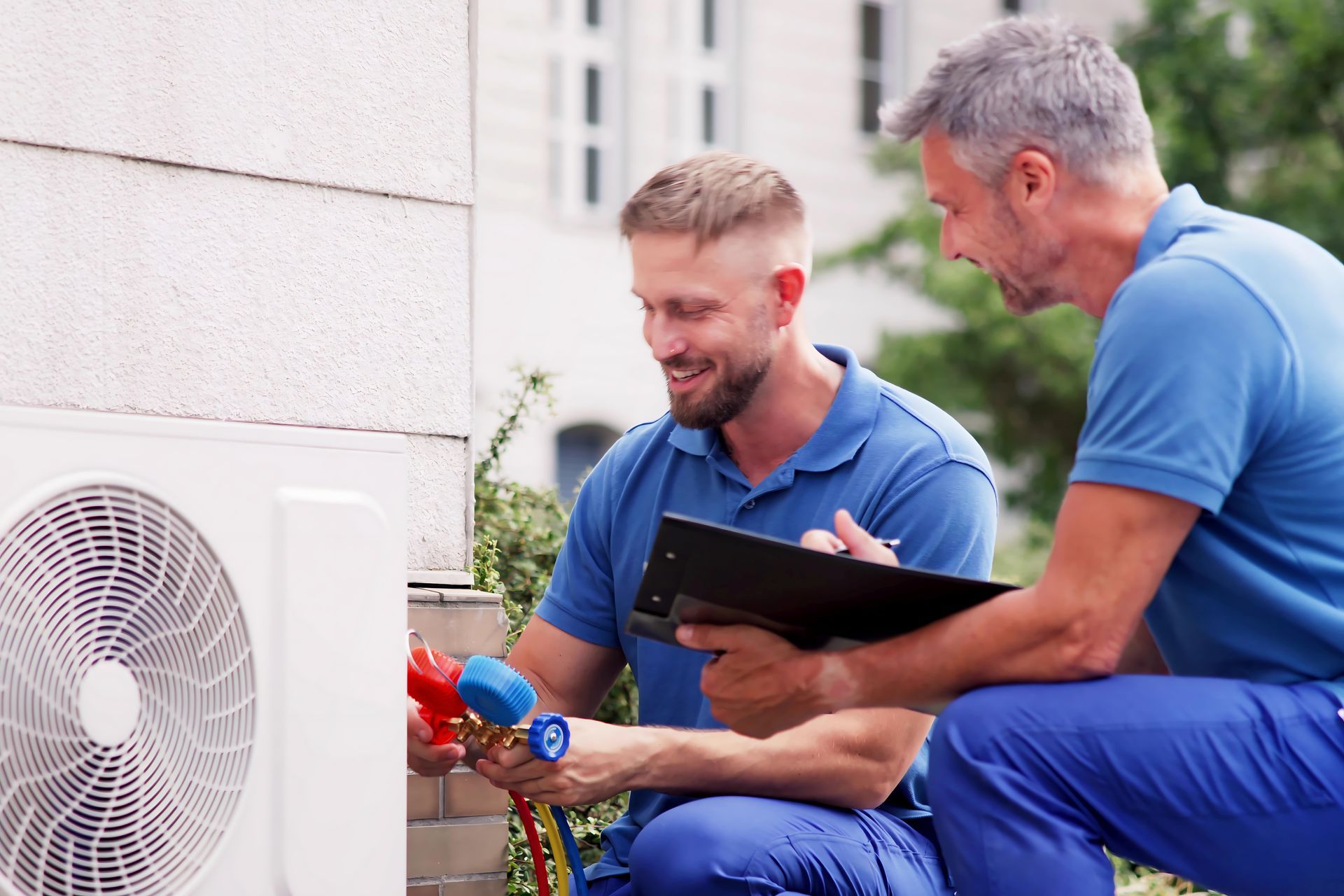 Two HVAC technicians inspecting an air conditioning unit. One kneels, connecting gauges. Outdoors.