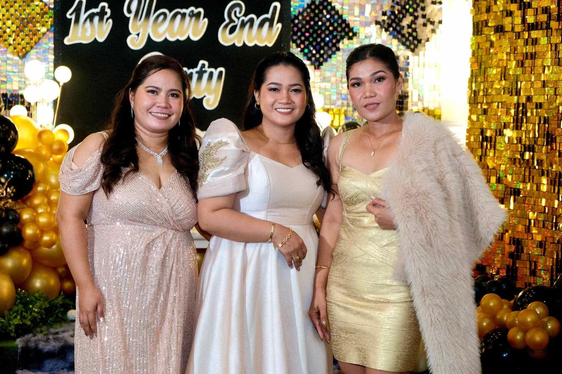 Three women at a party posing, smiling, and dressed in formal wear in front of gold decorations.