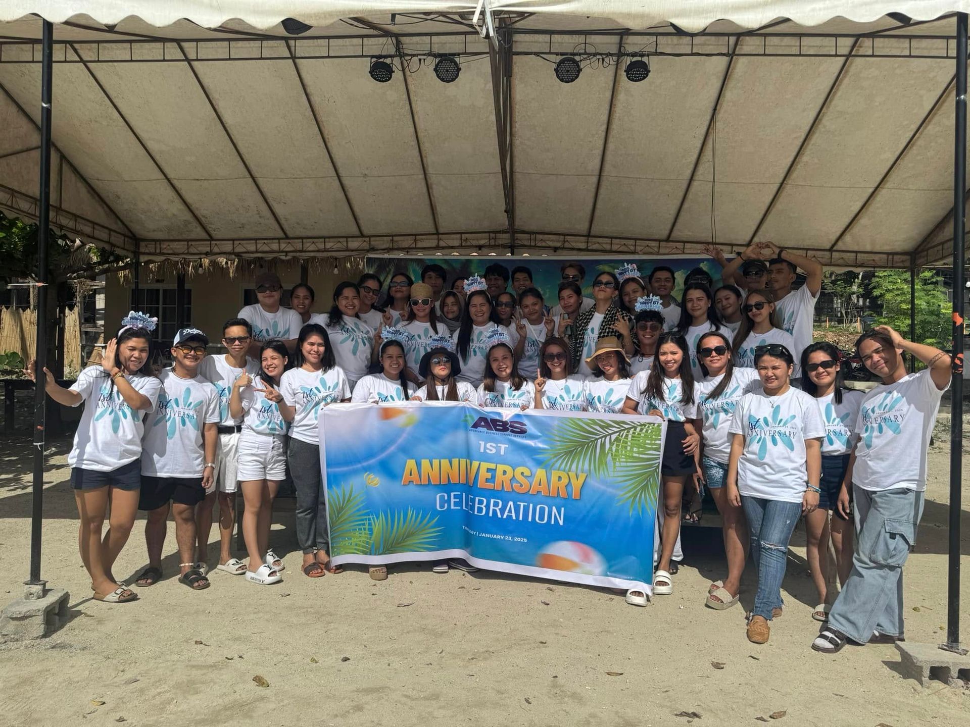 Group of people pose with a banner at an outdoor anniversary celebration. Most wear white shirts.