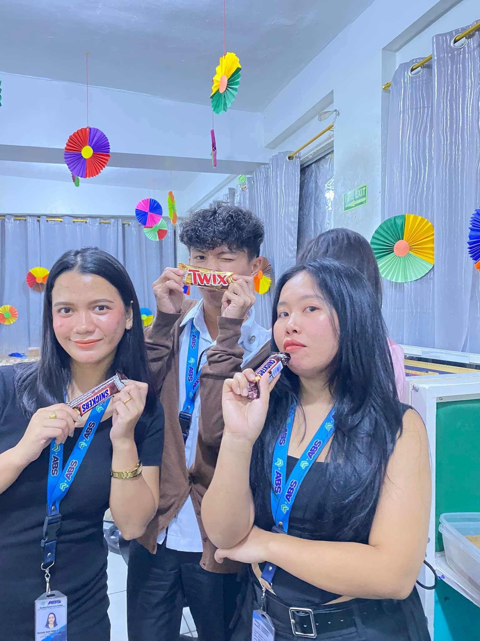 Three people holding candy bars, smiling. Room decorated with paper fans.