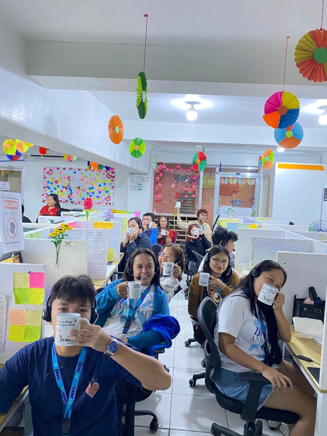 People in an office holding mugs, smiling. Cubicles, colorful decorations overhead, bright lights.