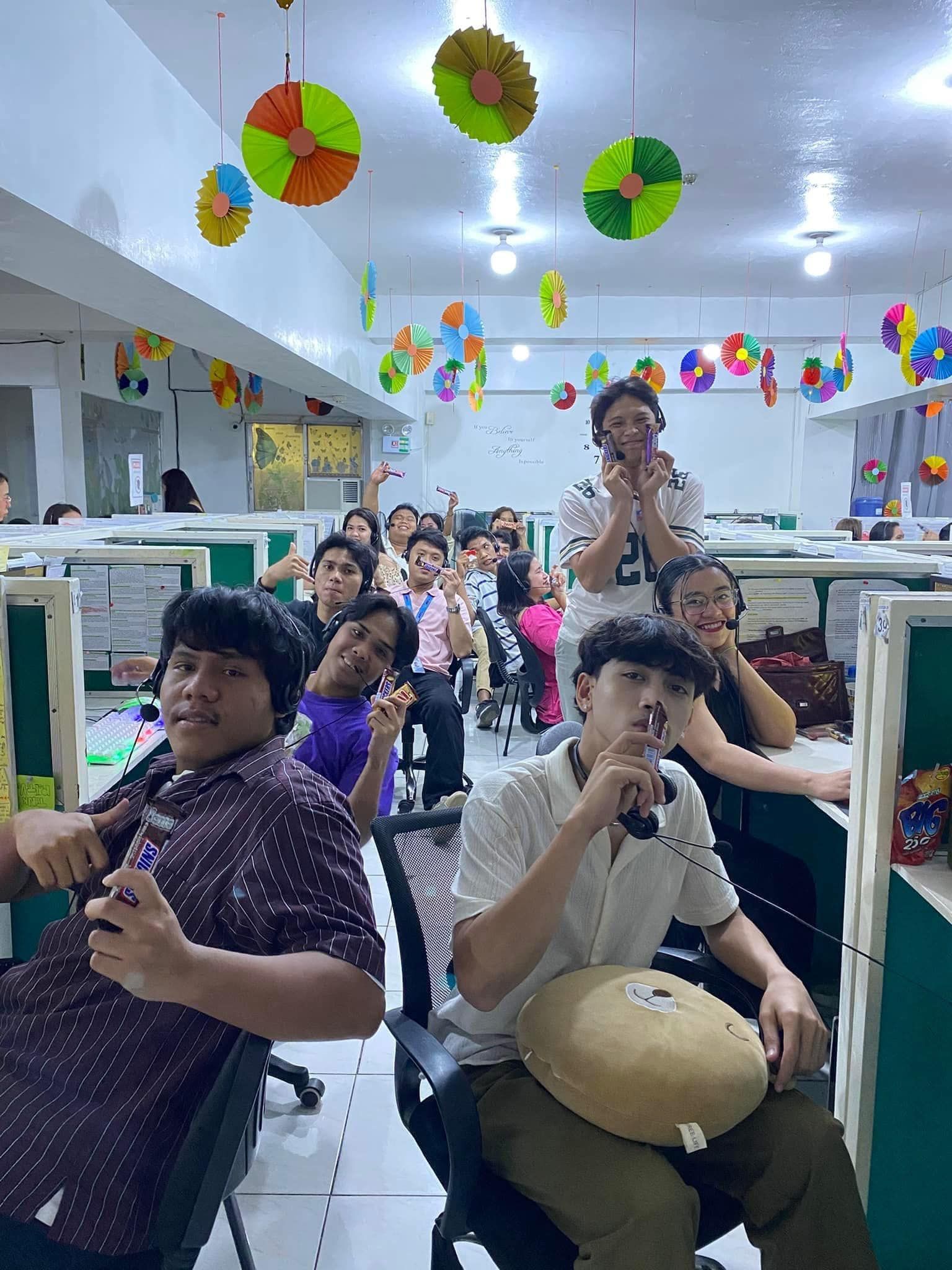Group of people in an office with colorful decorations, posing and smiling, some holding phones.