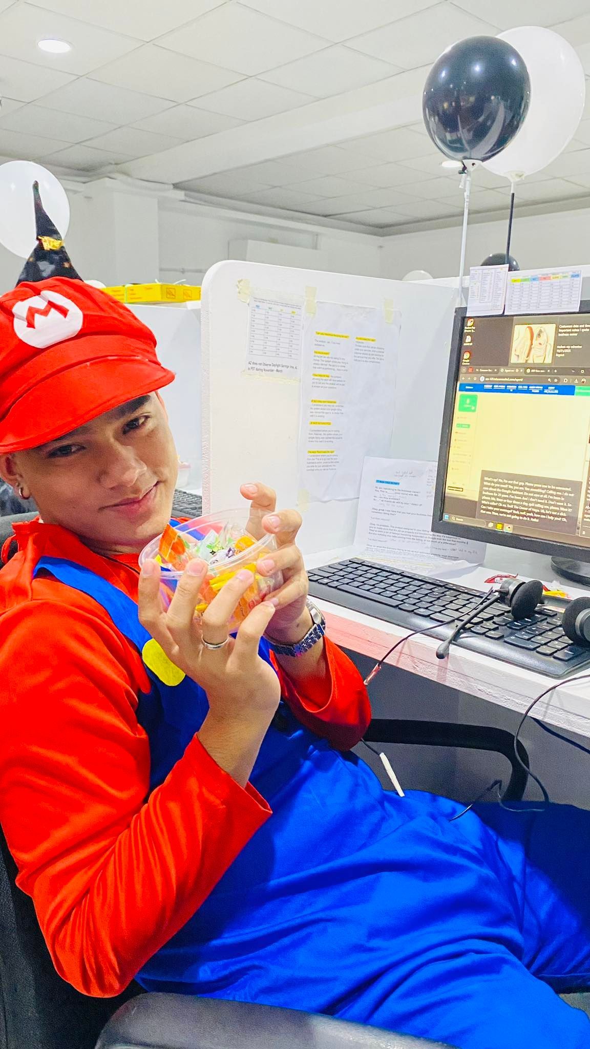 Person in Mario costume holding a Rubik's cube, at a desk with a computer and balloons.
