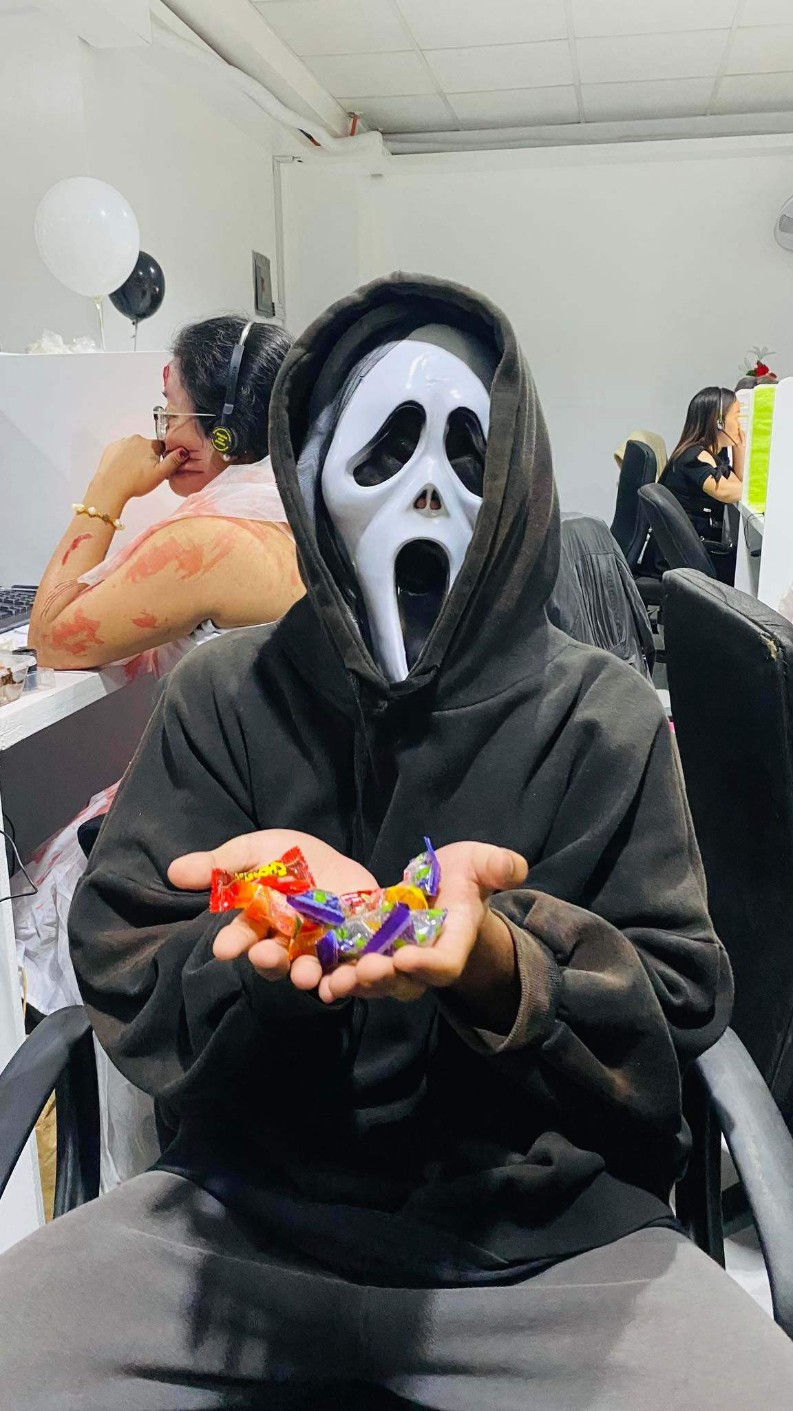 Person in Scream mask holding candy, seated. Halloween scene with others in background.