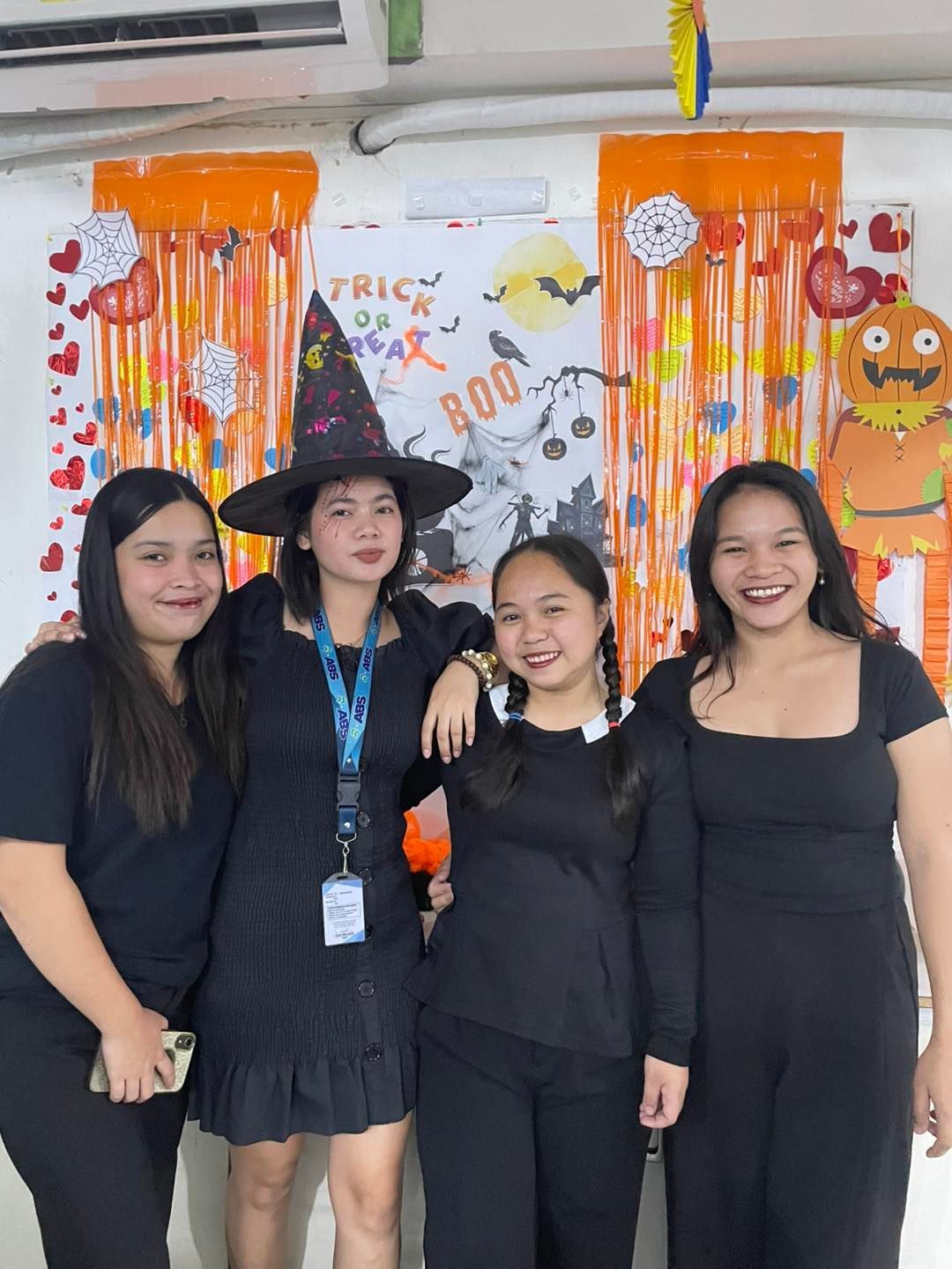 Four women in black outfits pose in front of Halloween decorations, including a witch hat and 