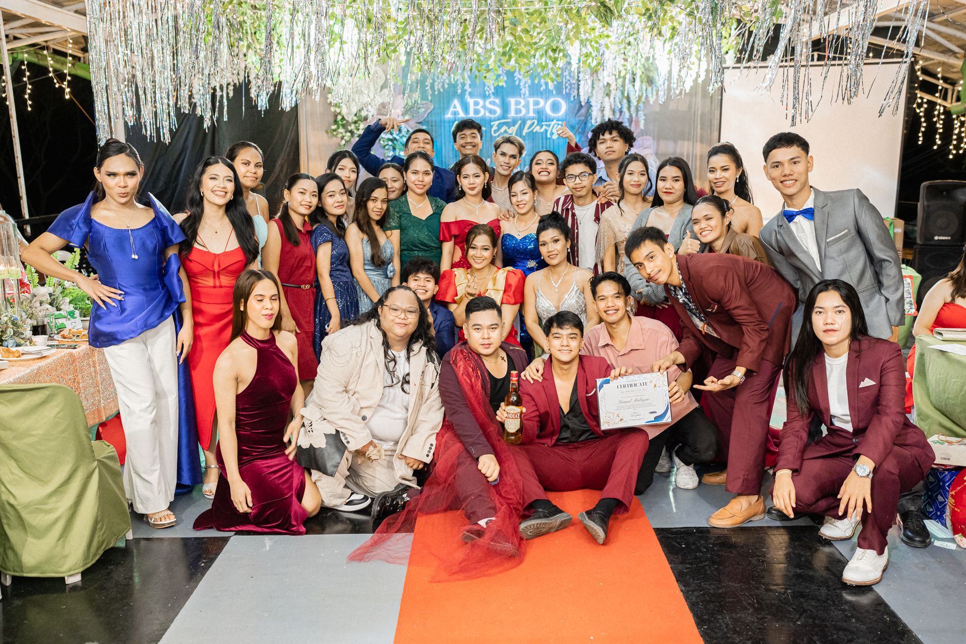 Group of people posing at an event. Some in formal attire, red carpet, colorful background, festive mood.