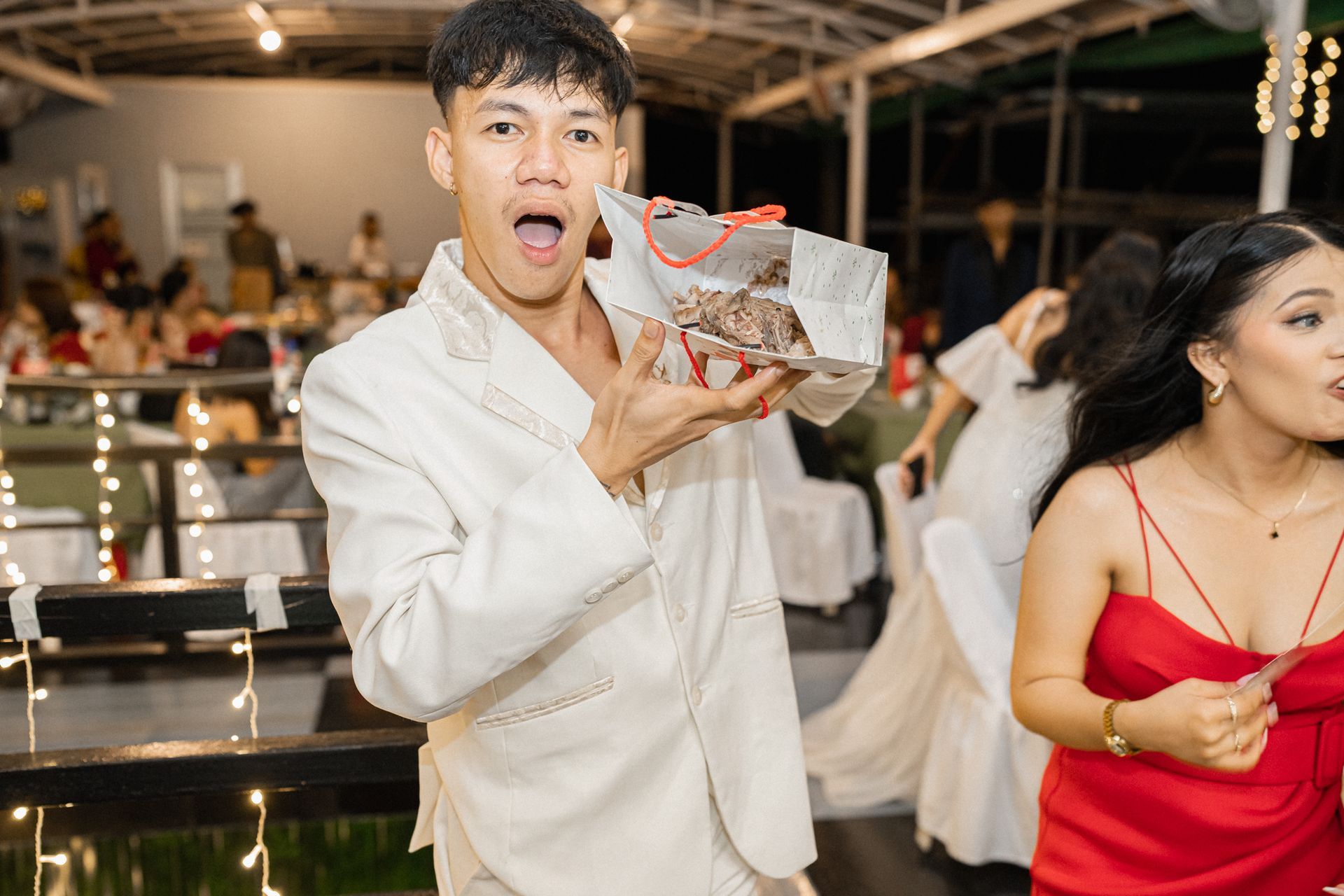 Man in white suit excitedly holding open a gift bag with dessert, party setting.