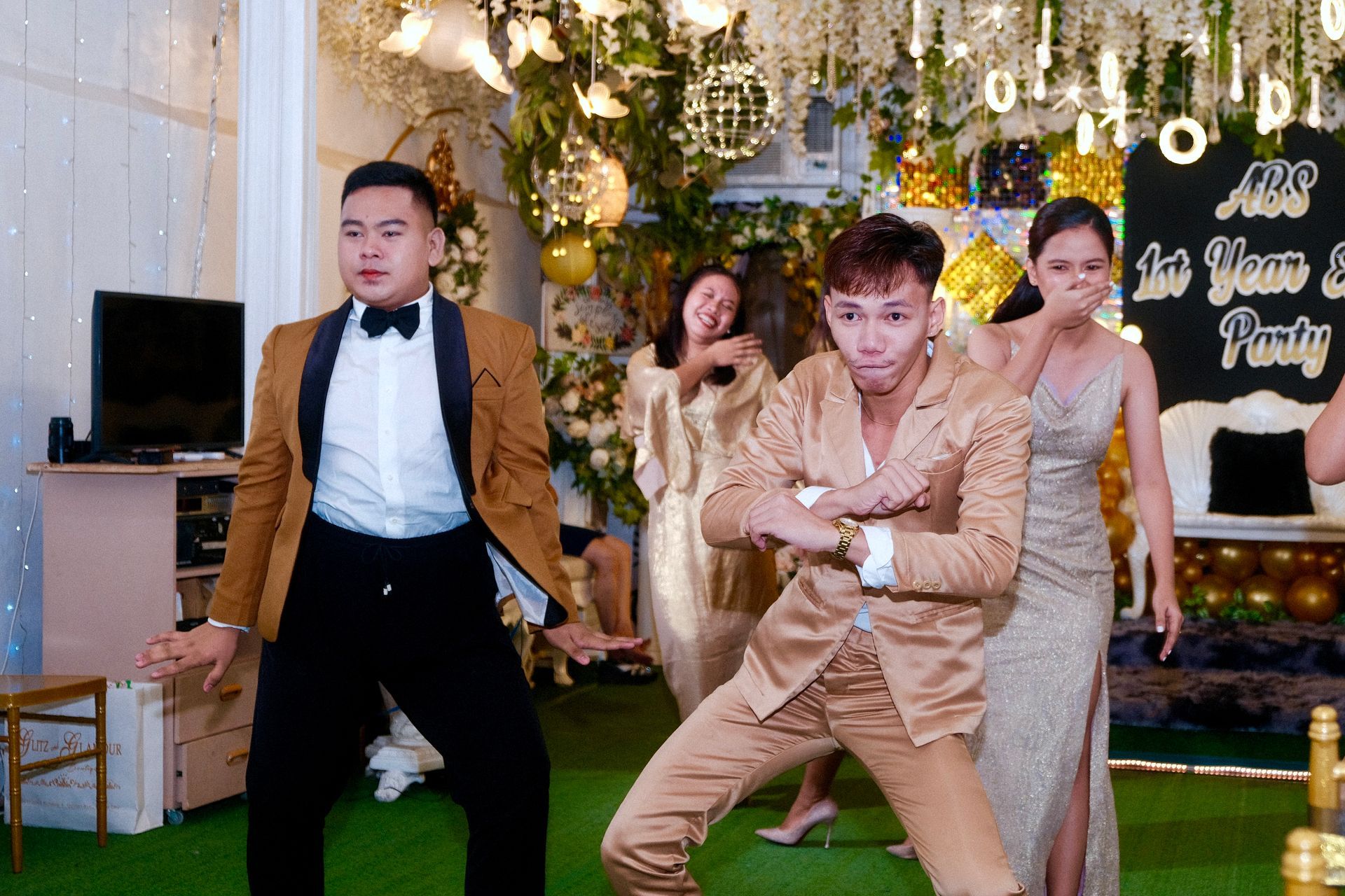 People dancing at a party with gold and white decorations. One man in a gold suit dances, others in formal attire.
