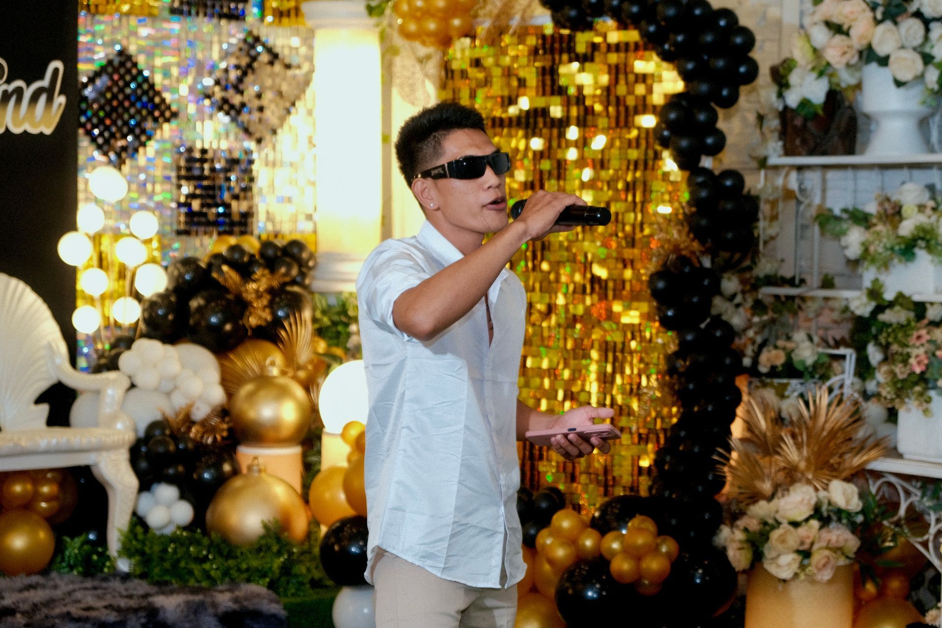 Man in sunglasses sings into a microphone at a party with gold and black decorations.