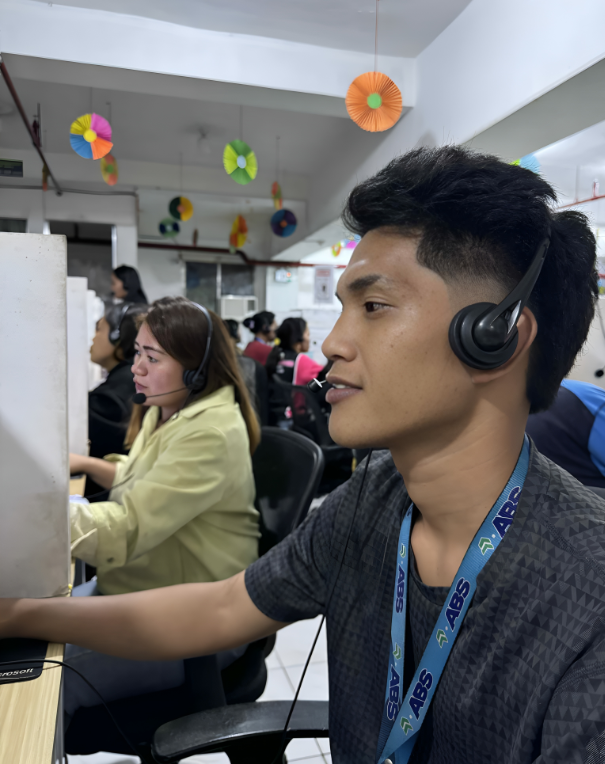 Man wearing headset in a call center, smiling. Other workers in background, colorful decorations.