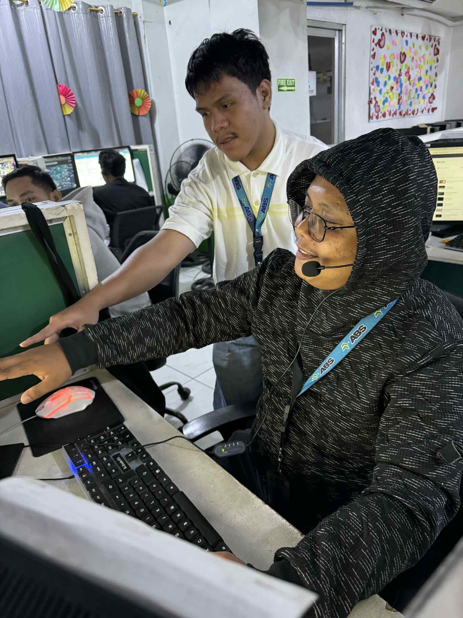 Man assisting another person at a computer. Indoor office setting, bright lighting. They are looking at the screen.