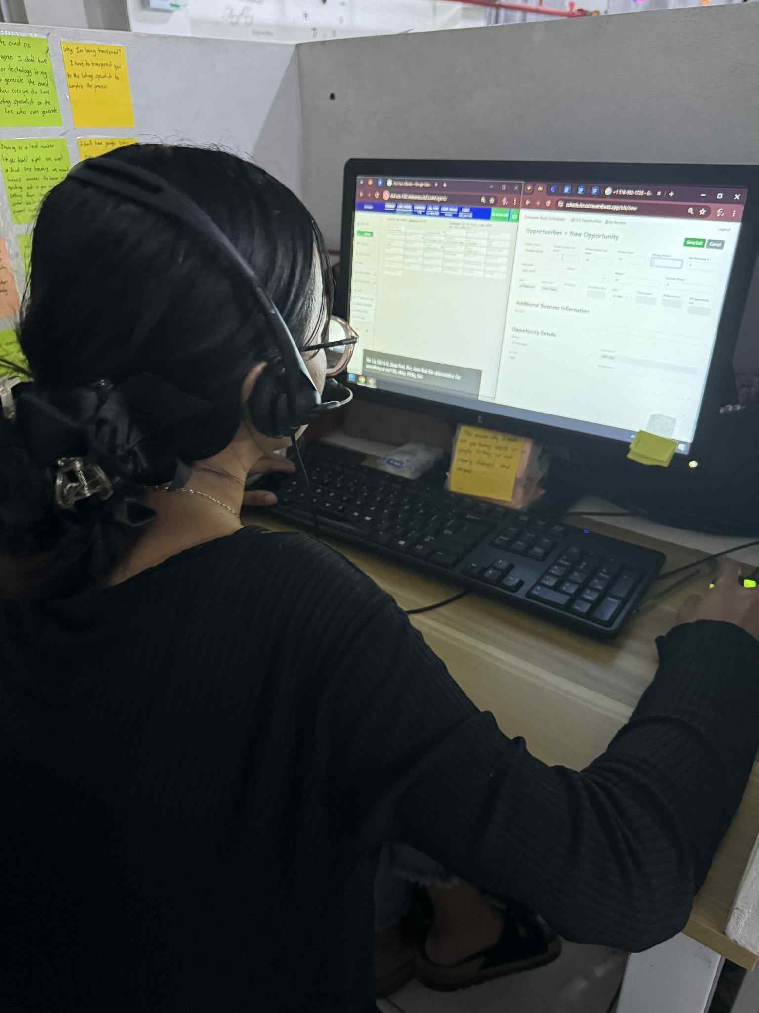Person using a computer, wearing a headset, in an office. Black shirt, dark hair, focused on the screen.