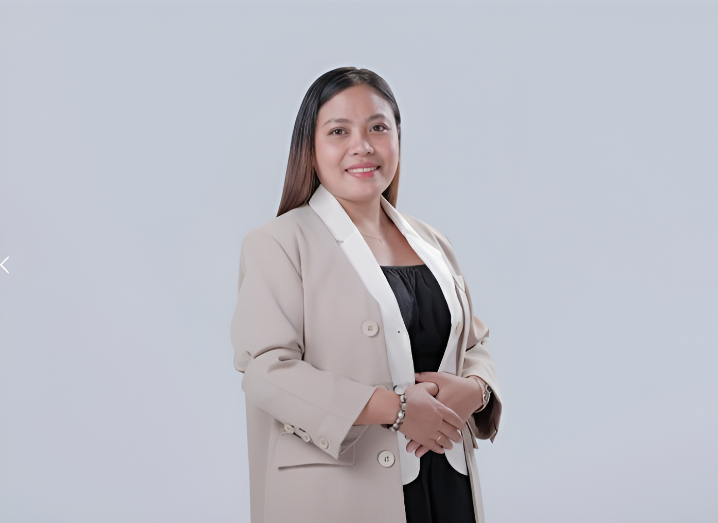 Woman in a beige blazer and black top smiles, hands clasped, against a white backdrop.